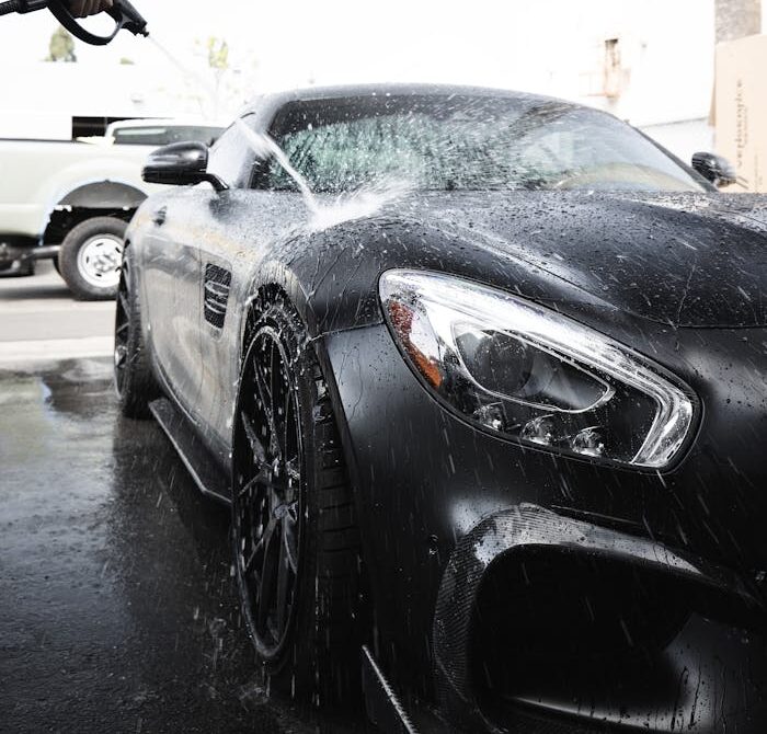 A luxury black car being washed with a pressure hose on a sunny day.