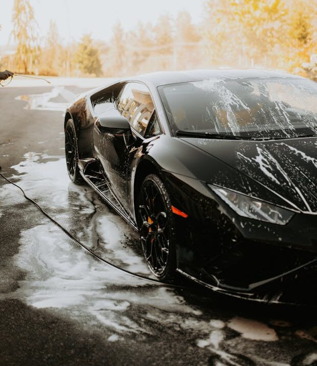 A man washing a sleek black Lamborghini sports car with soap suds outdoors.